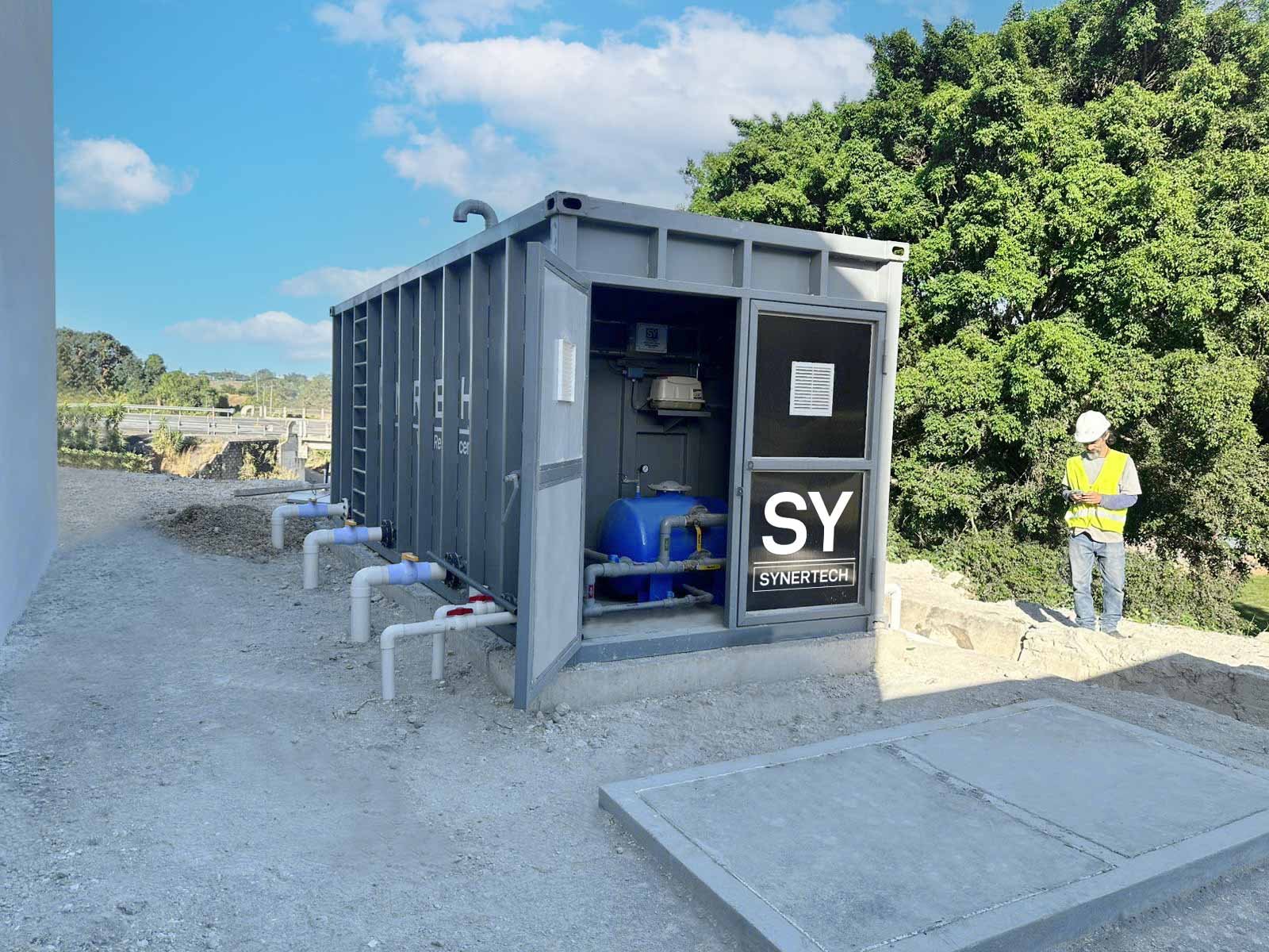 Técnico inspeccionando una planta de tratamiento de aguas residuales modular de color verde marca BIOMAX SYNERTECH, que incluye módulos de pretratamiento, decantación, DAF y oxidación avanzada. La planta está ubicada en un entorno industrial con techo alto y tuberías visibles. - ver más información